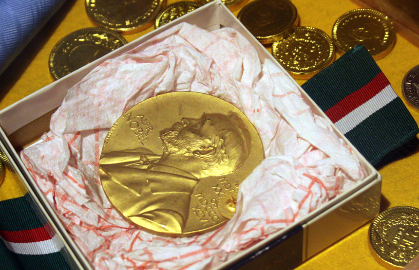 Złoty medal Nobla w ozdobnym pudełku, otoczony kolorowymi monetami.