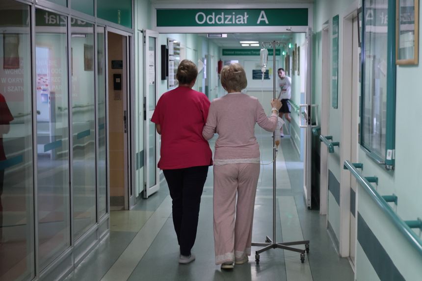 Dwie osoby idą korytarzem szpitalnym, jedna z wózkiem na kroplówkę, druga w różowej koszulce.