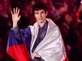 Młody mężczyzna w ciemnej koszuli, machający ręką, owinięty flagą. Tło z tłumem i światłami.