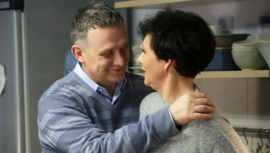 Para w uścisku w kuchni, uśmiechają się do siebie. W tle półki z naczyniami.