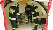 Czterech strażaków w mundurach ratunkowych transportuje poszkodowanego na noszach w namiocie.