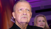 Mężczyzna zrzuca poważne spojrzenie, w tle widoczna kobieta i delikatne światło.