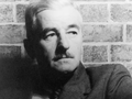 Mężczyzna z wąsami w szarym garniturze, siedzący przed ceglanym murem. Czarnobiałe zdjęcie.