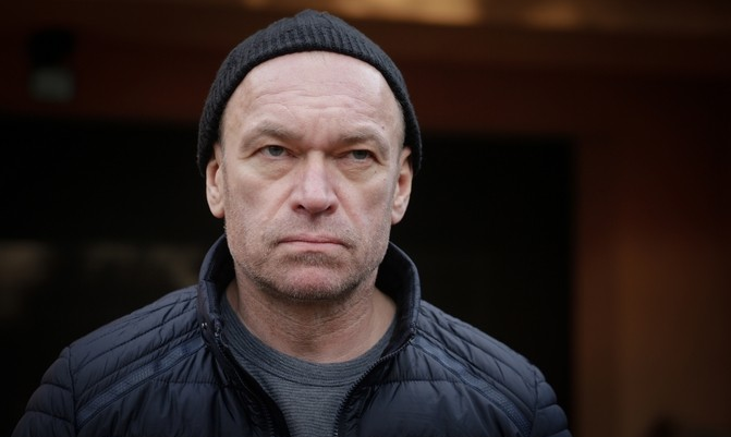 Mężczyzna w czarnej beanie i kurtce, z poważnym wyrazem twarzy, stoi na tle ciemnego tła.