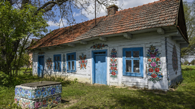 Tradycyjny dom z białego drewna, zdobiony kolorowymi kwiatami, wśród zieleni i błękitu nieba.