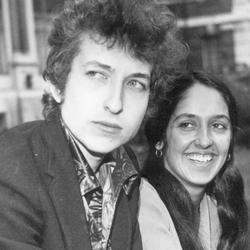 Bob Dylan i Joan Baez w 1965 roku. Młody mężczyzna z kręconymi włosami i uśmiechnięta kobieta na tle budynków.