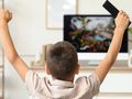 Chłopiec z uniesioną ręką i pilotem, oglądający telewizor w jasnym wnętrzu.