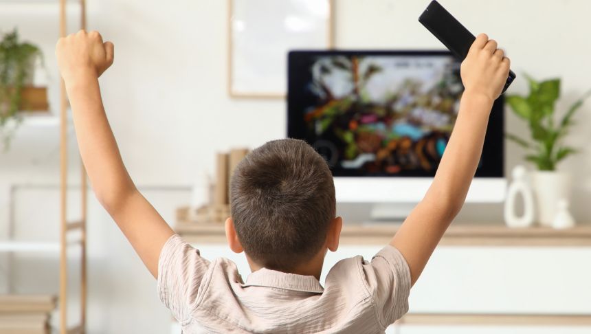 Chłopiec z uniesioną ręką i pilotem, oglądający telewizor w jasnym wnętrzu.