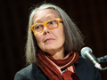 Anne Carson. Na zdjęciu widać kobietę z szarymi włosami, w okularach i z chustą na szyi, siedzącą przy mikrofonie.