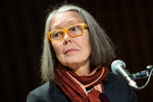 Anne Carson. Na zdjęciu widać kobietę z szarymi włosami, w okularach i z chustą na szyi, siedzącą przy mikrofonie.