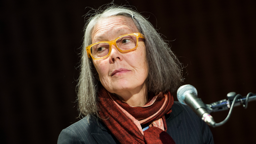 Anne Carson. Na zdjęciu widać kobietę z szarymi włosami, w okularach i z chustą na szyi, siedzącą przy mikrofonie.