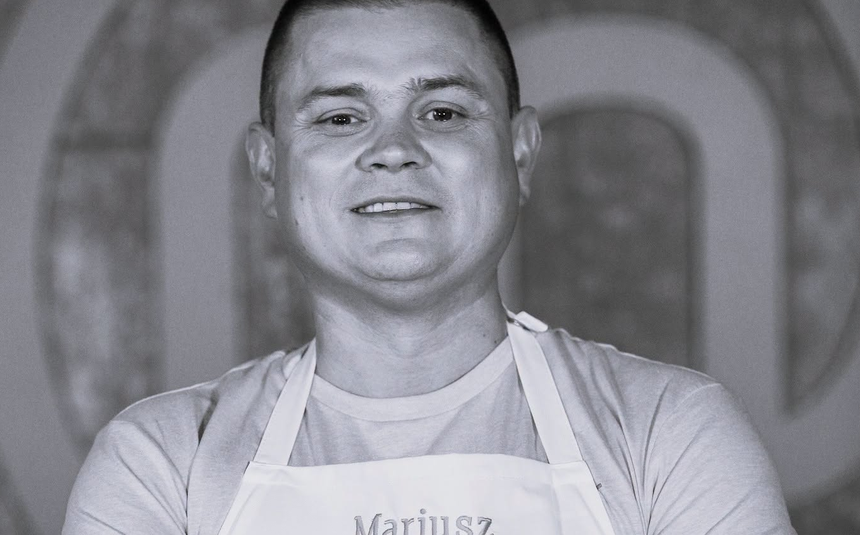 Mariusz Komenda w jasnej koszulce i fartuchu kuchennym, uśmiechnięty, w tle rozmyte kształty.