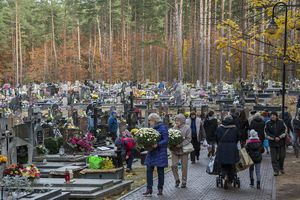 Widok na cmentarz z ludźmi niosącymi kwiaty, drzewami w tle, w jesiennej scenerii.