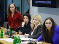 Cztery kobiety w garniturach na konferencji, jedna w czerwonym swetrze, przy stole z mikrofonami.