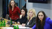 Cztery kobiety w garniturach na konferencji, jedna w czerwonym swetrze, przy stole z mikrofonami.