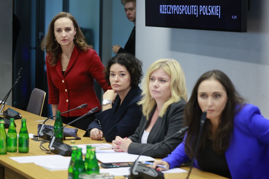 Cztery kobiety w garniturach na konferencji, jedna w czerwonym swetrze, przy stole z mikrofonami.
