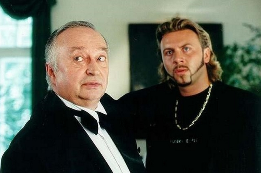 Na zdjęciu dwaj mężczyźni w eleganckich ubraniach; jeden z nich z wyrazem zaskoczenia.