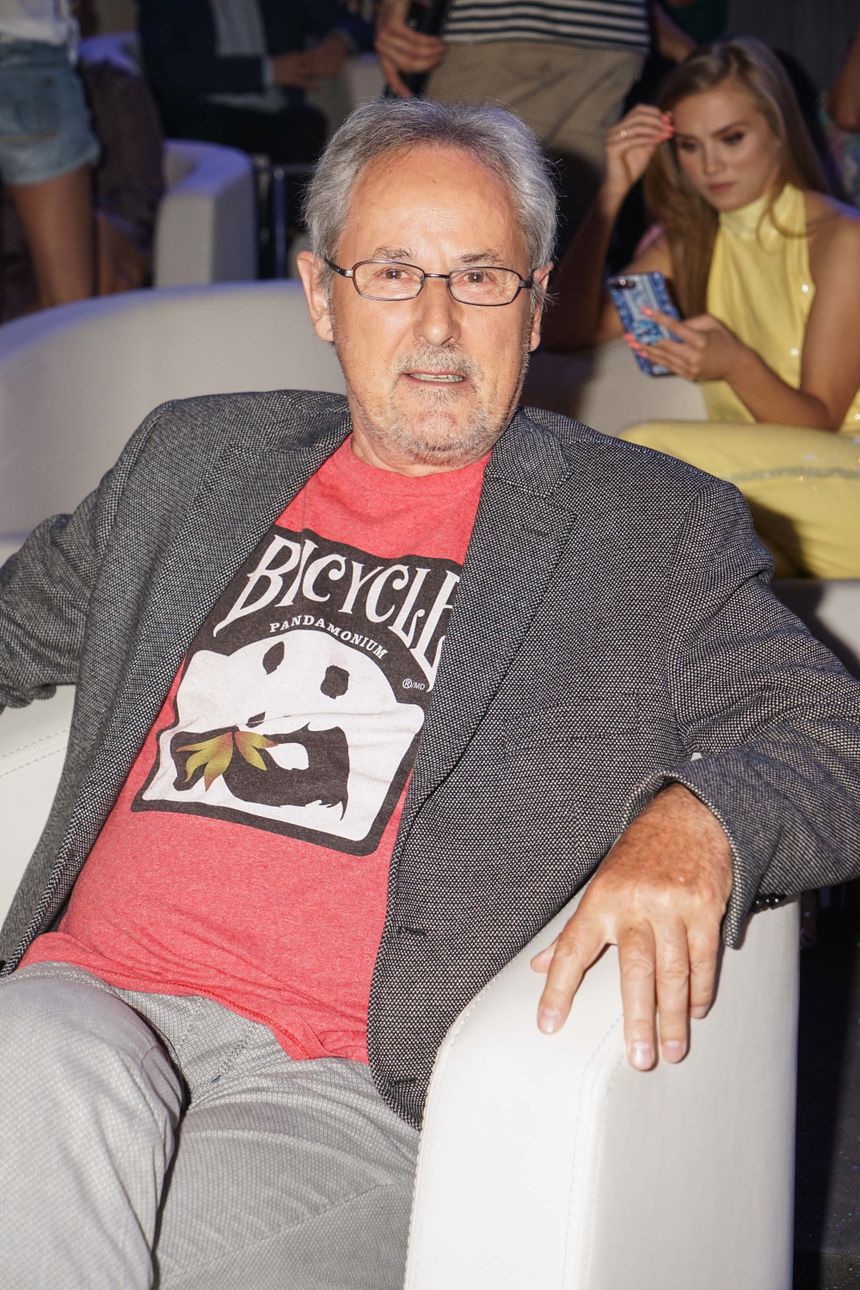 Mężczyzna w szarym garniturze i czerwonej koszulce siedzi na białej sofie, z poważnym wyrazem twarzy.