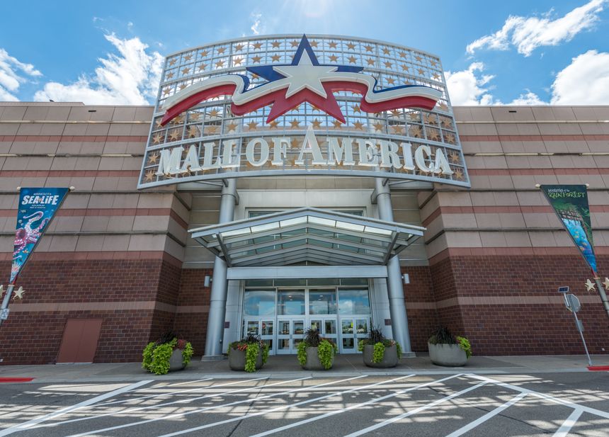 Wejście do Mall of America z dużym napisem i roślinnością przy drzwiach. Jasne niebo.