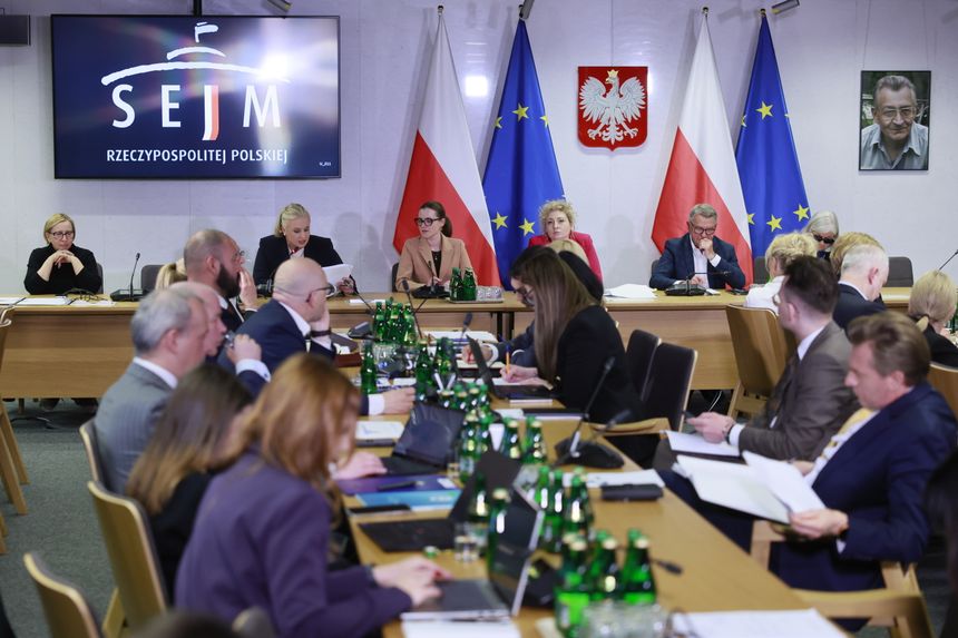 Posiedzenie Sejmu, uczestnicy siedzą przy stole, w tle flagi Polski i UE, logo Sejmu.