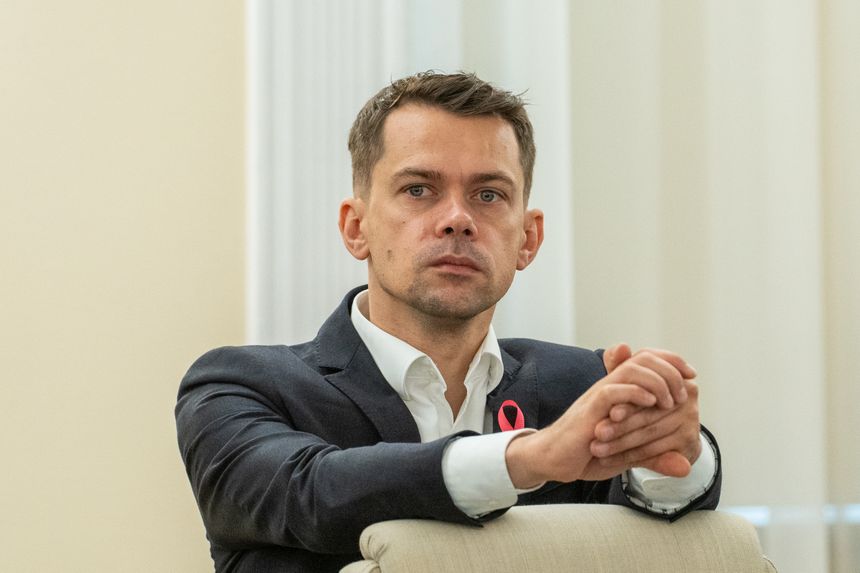 Mężczyzna w garniturze, siedzący na kanapie, z zamyśloną miną i złożonymi rękami.