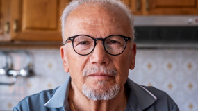 Mężczyzna w okularach z siwymi włosami i brodą, siedzi w kuchni z drewnianą szafką w tle.