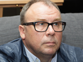 Mężczyzna w okularach, z zamyśloną miną, w niebieskiej kurtce, siedzi na szarej kanapie.