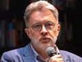 Mężczyzna z siwymi włosami i okularami, trzyma mikrofon i mówi do publiczności.