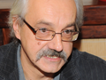 Mężczyzna z siwymi włosami i wąsami, w okularach, uśmiecha się w nieformalnym otoczeniu.
