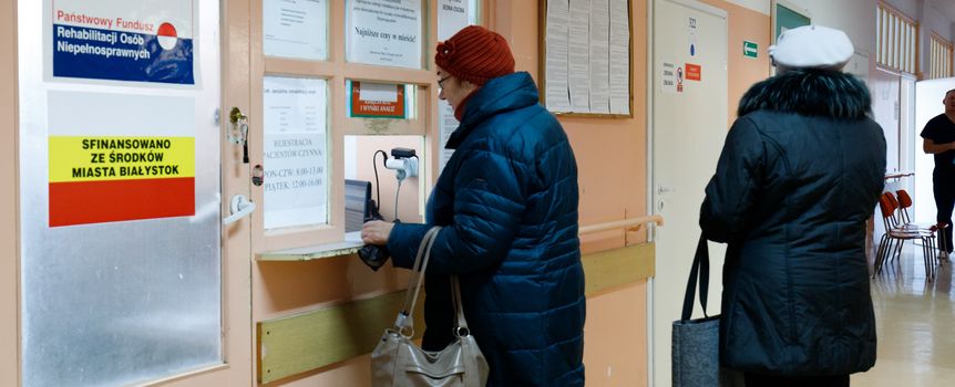 Na zdjęciu dwie kobiety w kurtkach stoją przy oknie rejestracyjnym w przychodni.