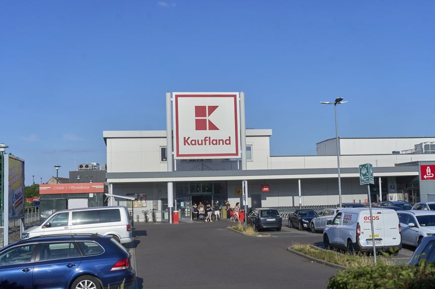 Widok na sklep Kaufland z parkingiem, błękitne niebo w tle, klienci przy wejściu.