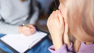 Osoba z różowymi włosami siedzi, trzymając dłońmi twarz, w tle piszący terapeuta.