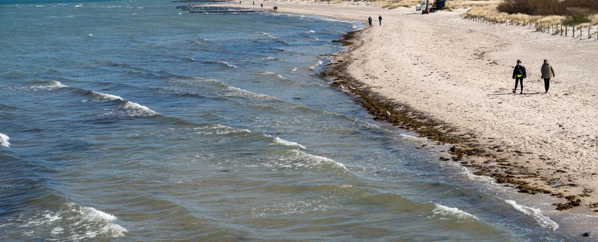 Plaża z falującym morzem, spacerujący ludzie wzdłuż brzegu i widok na wydmy w tle.