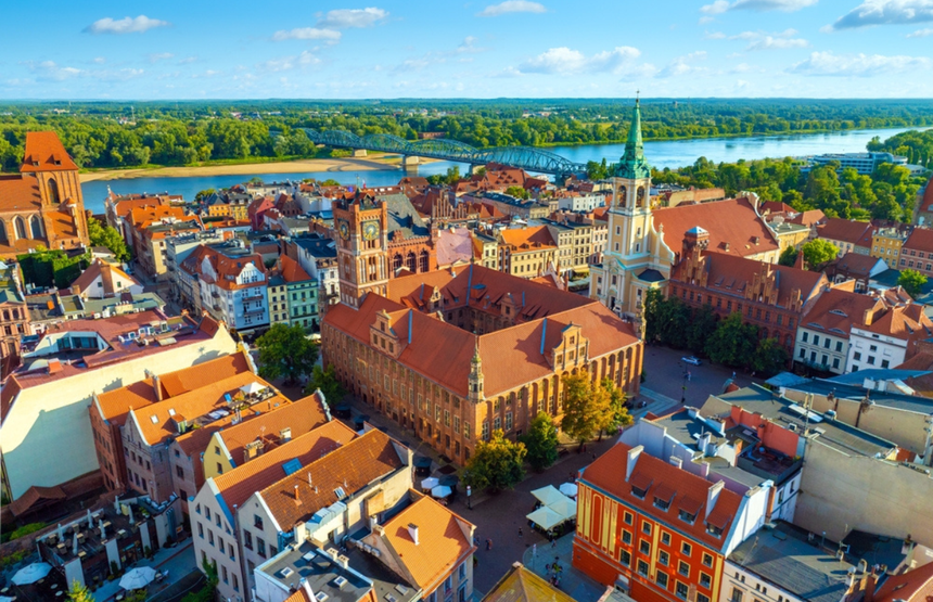 Widok na miasto z czerwonymi dachami, rzekę i zieleń w tle. Jasne niebo z chmurami.