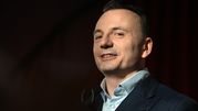 Mężczyzna w garniturze uśmiecha się, stojąc na tle ciemnej sali. 