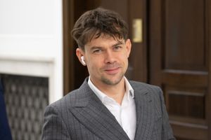 Mężczyzna w szarym garniturze z uśmiechem, w białej koszuli, słuchawki bezprzewodowe.
