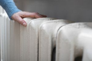 Ręka w niebieskim swetrze dotyka białego grzejnika. Wszystko w neutralnym oświetleniu.