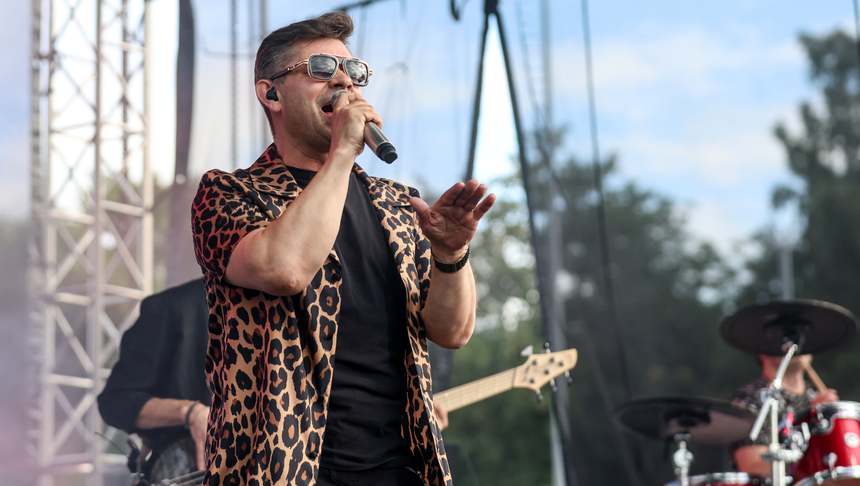 Mężczyzna w okularach słonecznych śpiewa na scenie, ubrany w kolorową koszulę w leoparda.