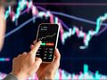 Osoba trzyma telefon, na którym wyświetlony jest wykres kursu Bitcoina. Tło to wykres finansowy.