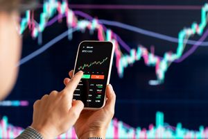 Osoba trzyma telefon, na którym wyświetlony jest wykres kursu Bitcoina. Tło to wykres finansowy.