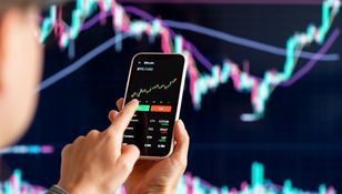 Osoba trzyma telefon, na którym wyświetlony jest wykres kursu Bitcoina. Tło to wykres finansowy.