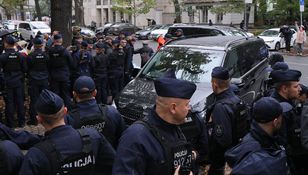 Grupa policjantów w mundurach, zabezpieczająca teren wokół czarnego minivana.