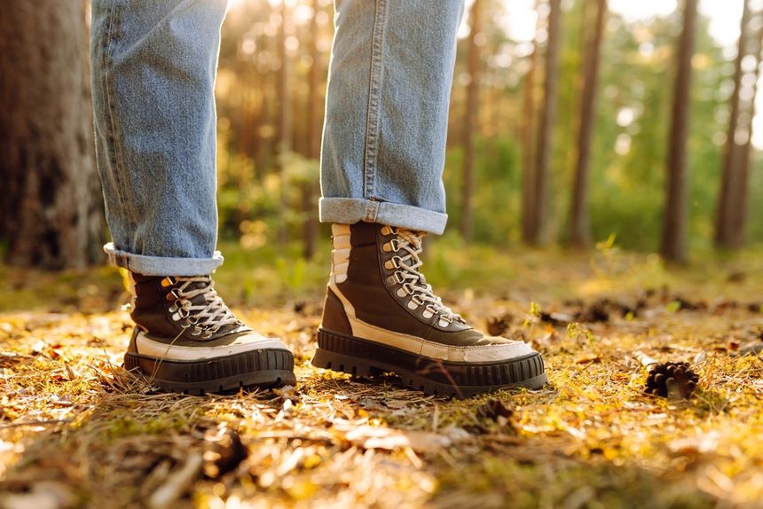 Osoba stoi na leśnej ścieżce w butach trekkingowych, otoczona drzewami i opadającymi liśćmi.