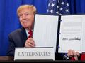 Mężczyzna w garniturze trzyma dokument, uśmiechając się. Za nim flaga USA.