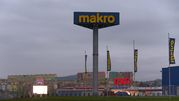 Widzok sklepy Makro i Tesco, w tle kolorowe bloki mieszkalne, szare niebo.