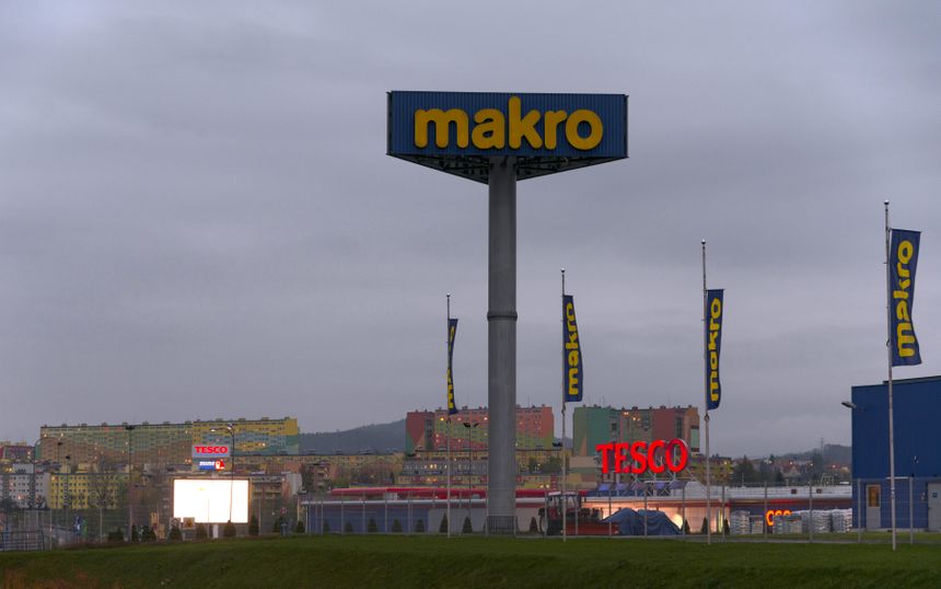 Widzok sklepy Makro i Tesco, w tle kolorowe bloki mieszkalne, szare niebo.