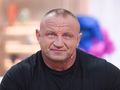 Mariusz Pudzianowski w czarnym t-shircie, z mikrofonem na uchu, uśmiecha się.
