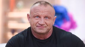 Mariusz Pudzianowski w czarnym t-shircie, z mikrofonem na uchu, uśmiecha się.