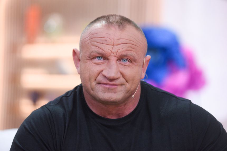 Mariusz Pudzianowski w czarnym t-shircie, z mikrofonem na uchu, uśmiecha się.