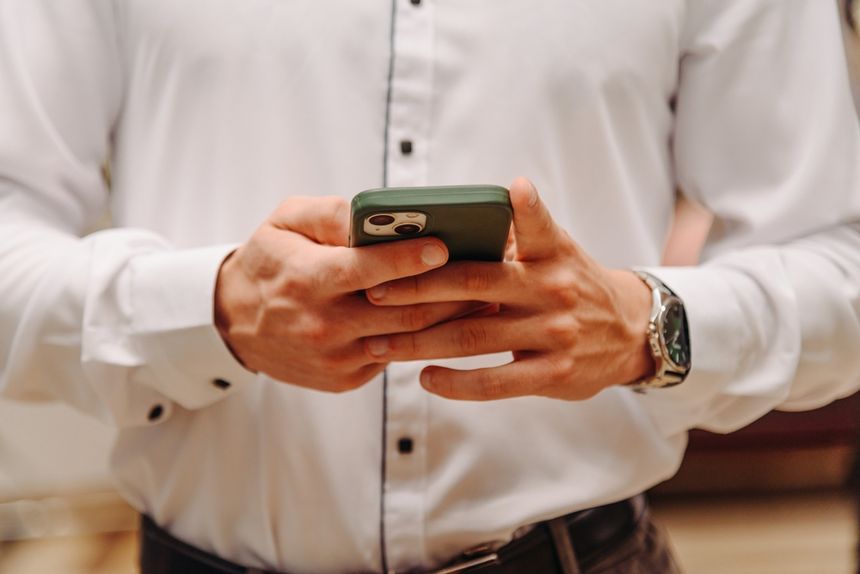 Mężczyzna w białej koszuli trzyma telefon komórkowy w dłoniach. Na nadgarstku ma zegarek.